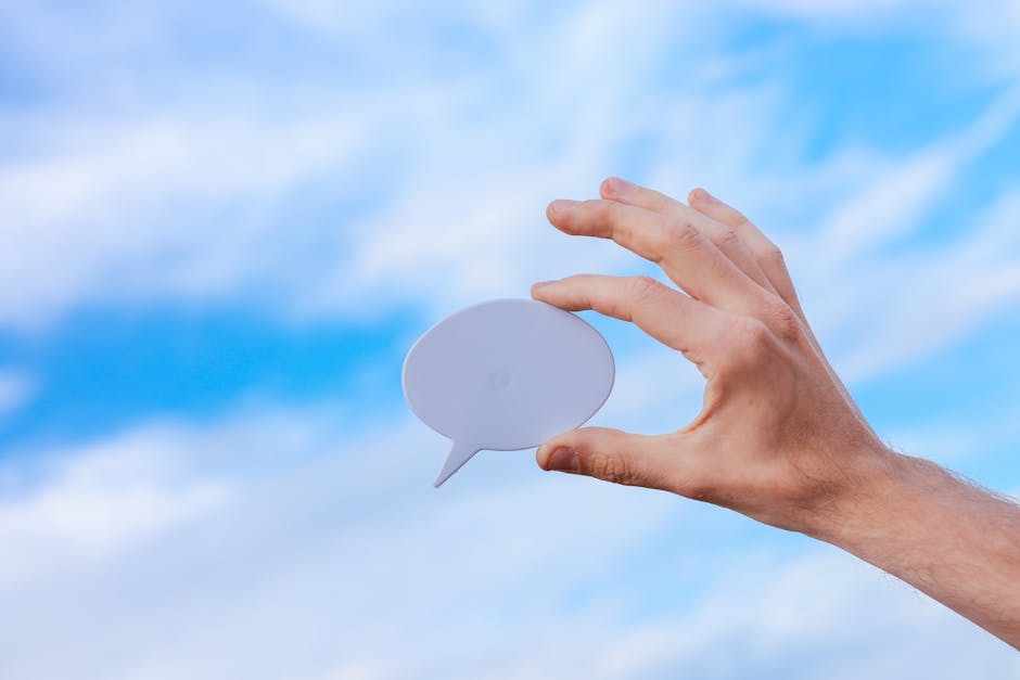 Hand holding a speech bubble against a vibrant blue sky with scattered clouds.