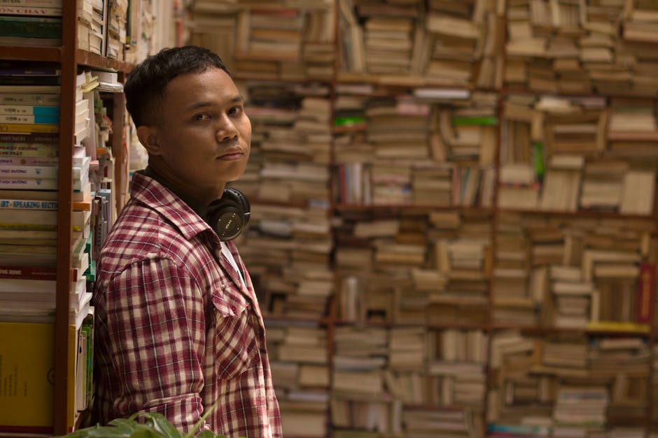 Portrait of a man in a library with shelves full of books, wearing headphones and plaid shirt.
